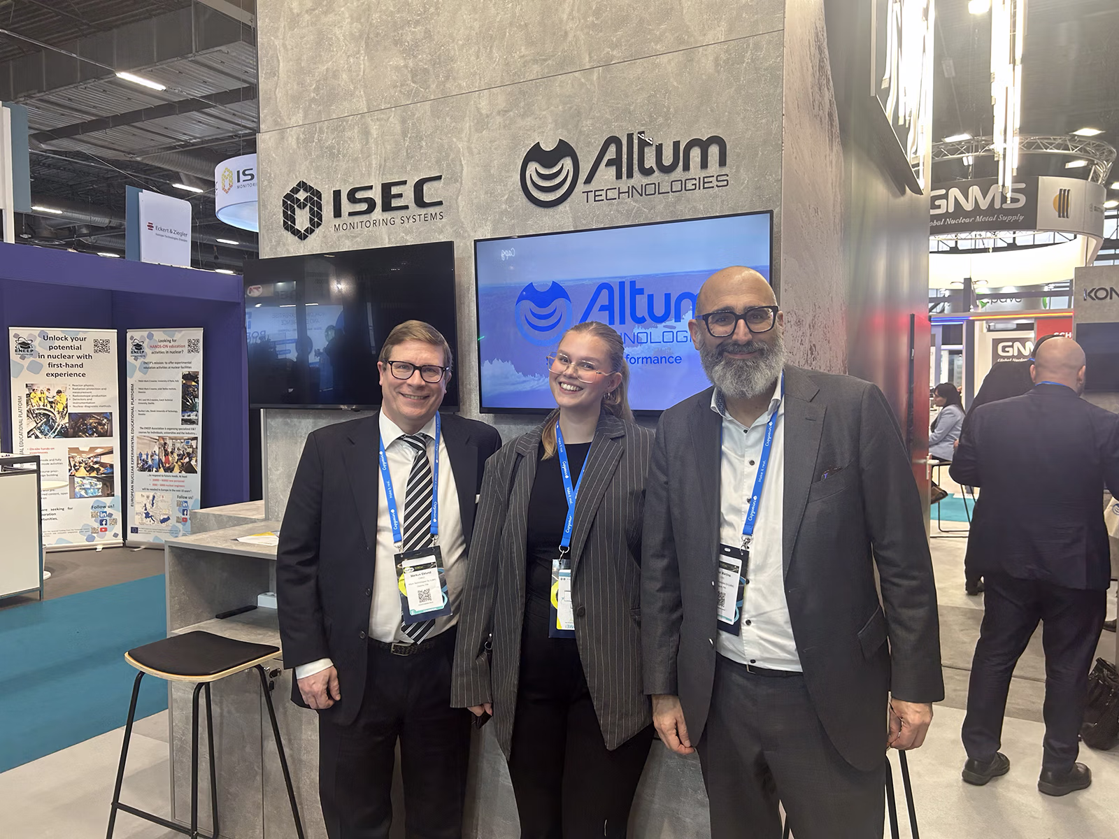 Three people standing in front of a exhibition booth with the words Altum Technologies written on it.